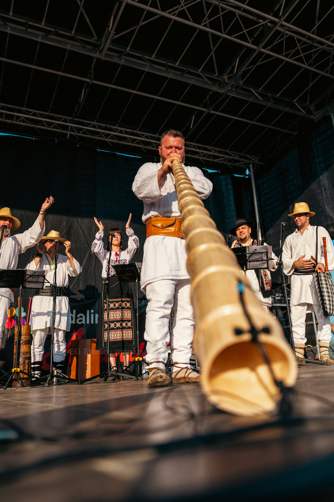 Low-angle view of a folk musician playing a long wooden horn directly toward the camera.