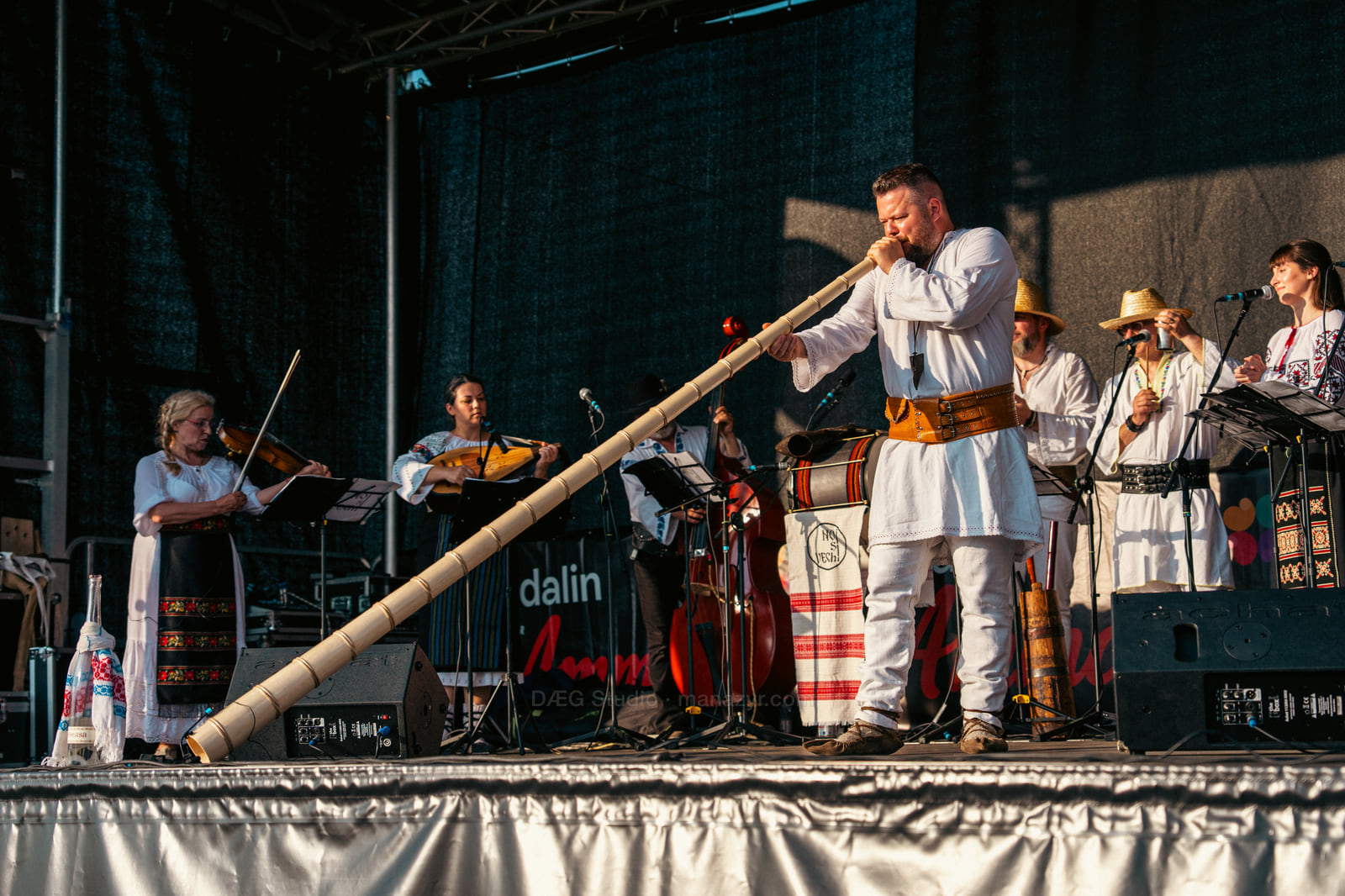 Folk ensemble performs in traditional clothing as a musician plays a long wooden horn across the stage.