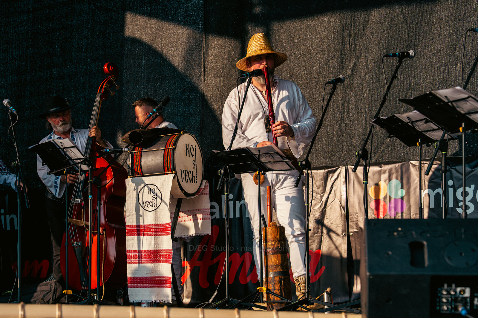 Folk musicians perform among double bass, drum and traditional wind instruments in angled evening light.