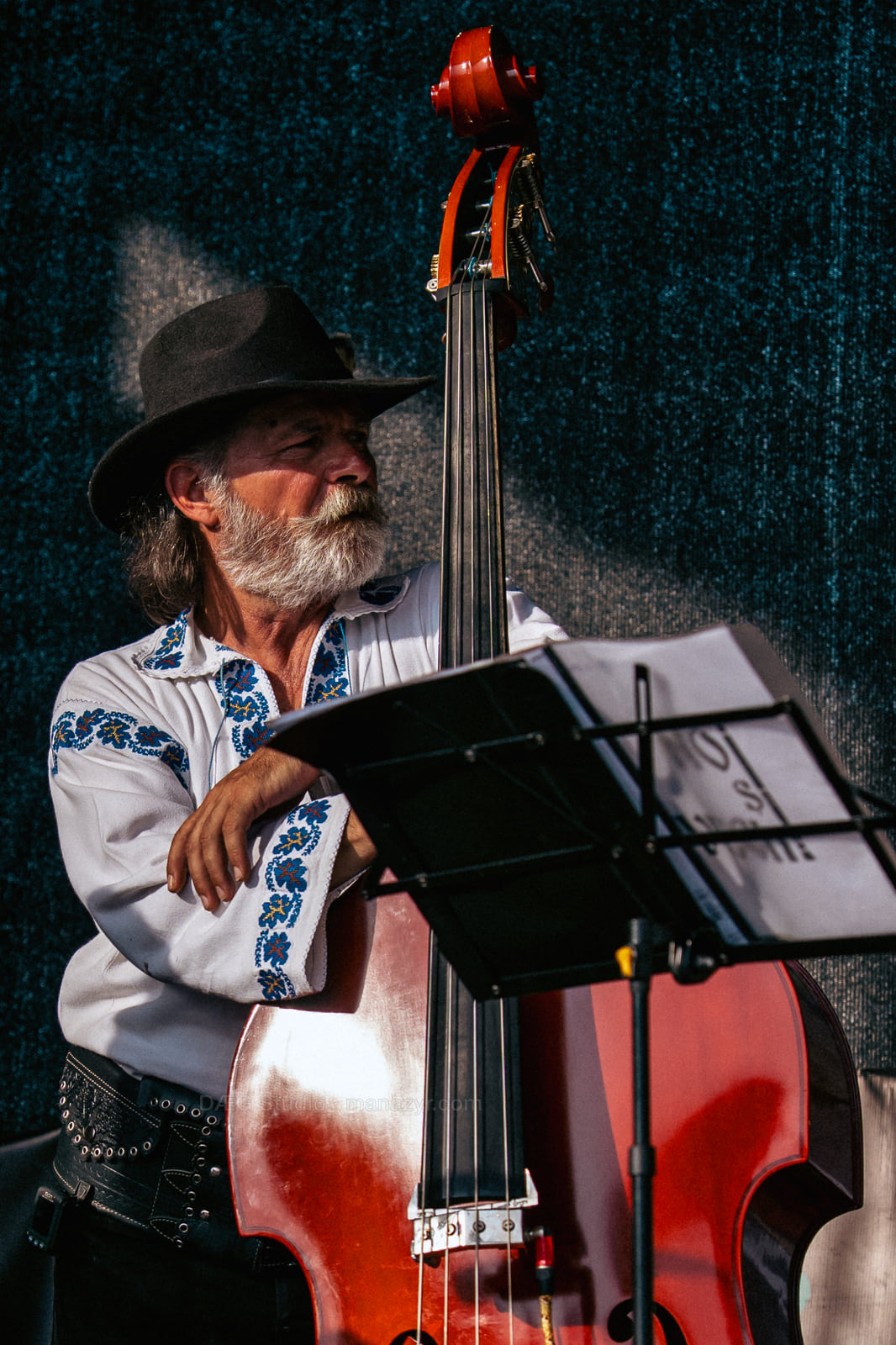 Older double bassist in an embroidered white shirt and black hat pauses in warm stage light.