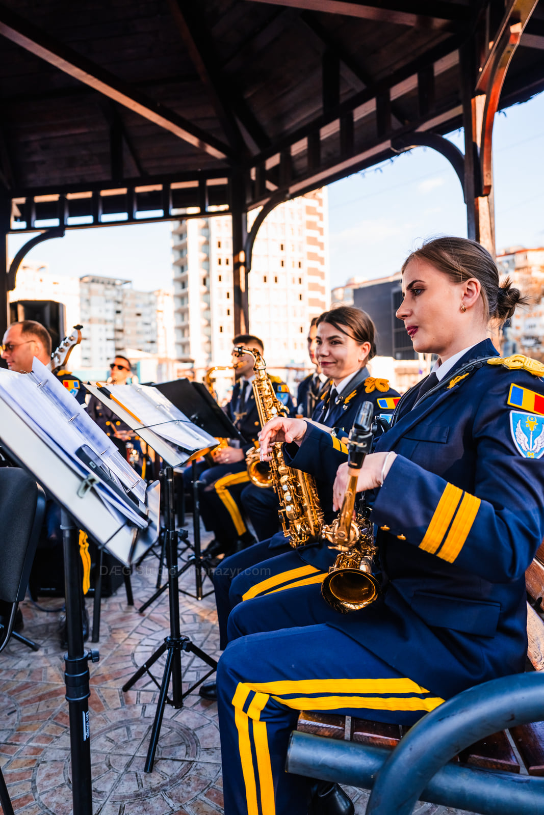 Uniformed saxophonists perform in a seated row beneath an outdoor pavilion.