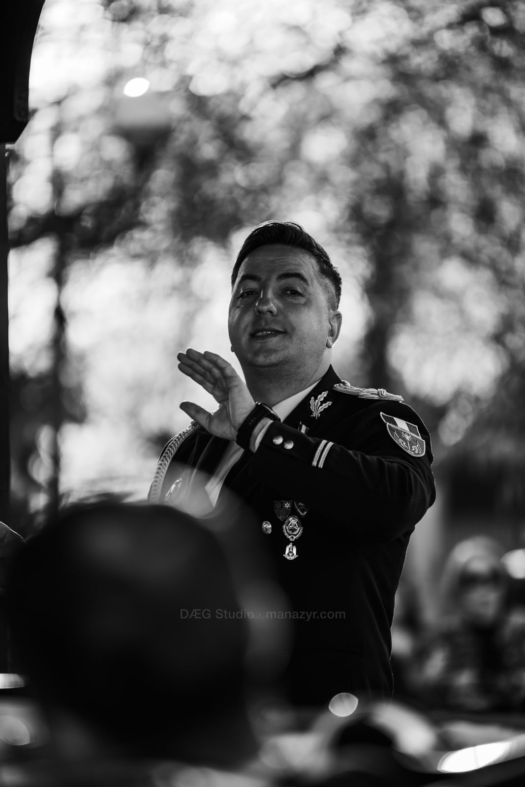 Black-and-white portrait of the conductor gesturing above blurred instruments and music stands.