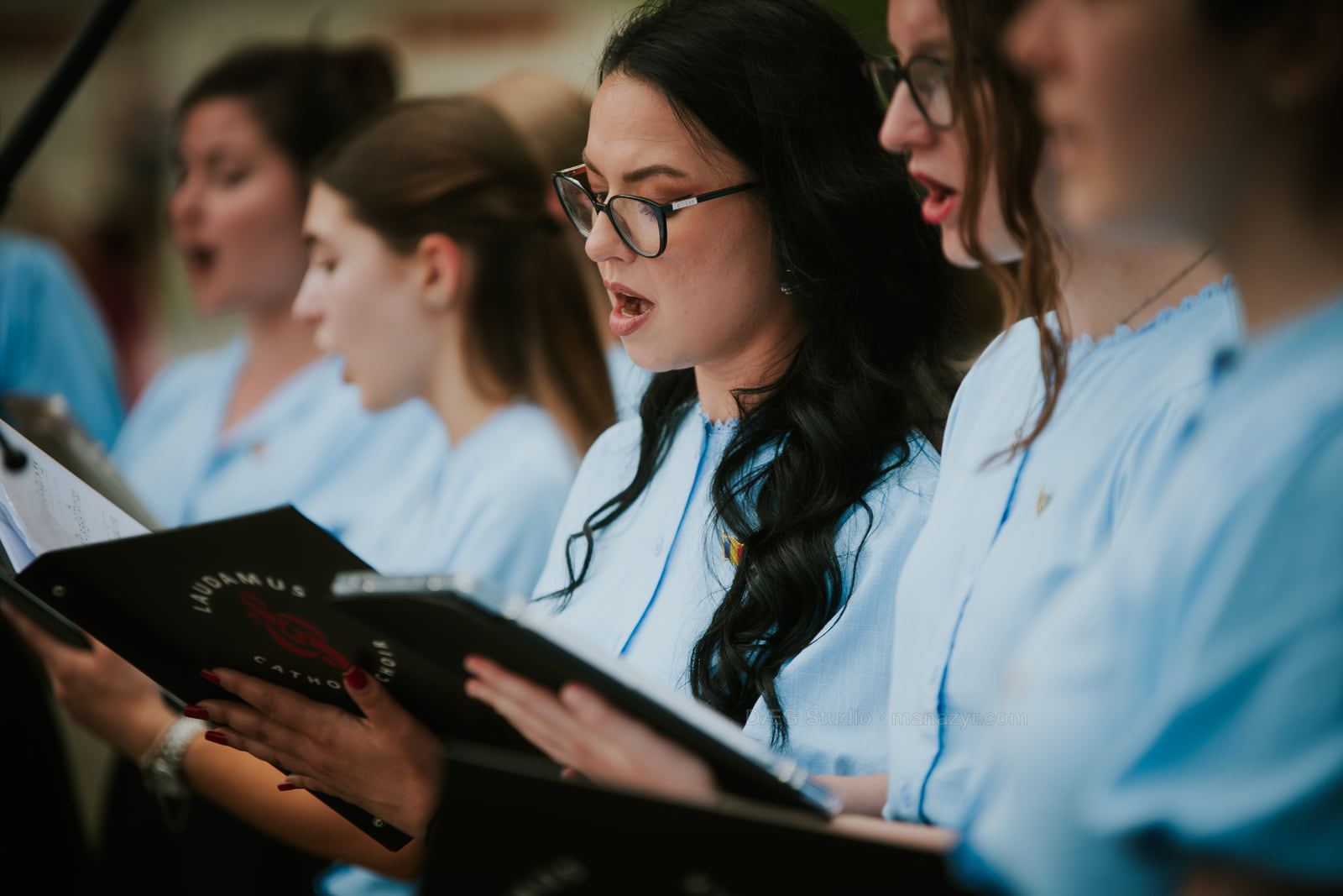 Women’s choir in pale blue blouses sings from open black music folders.