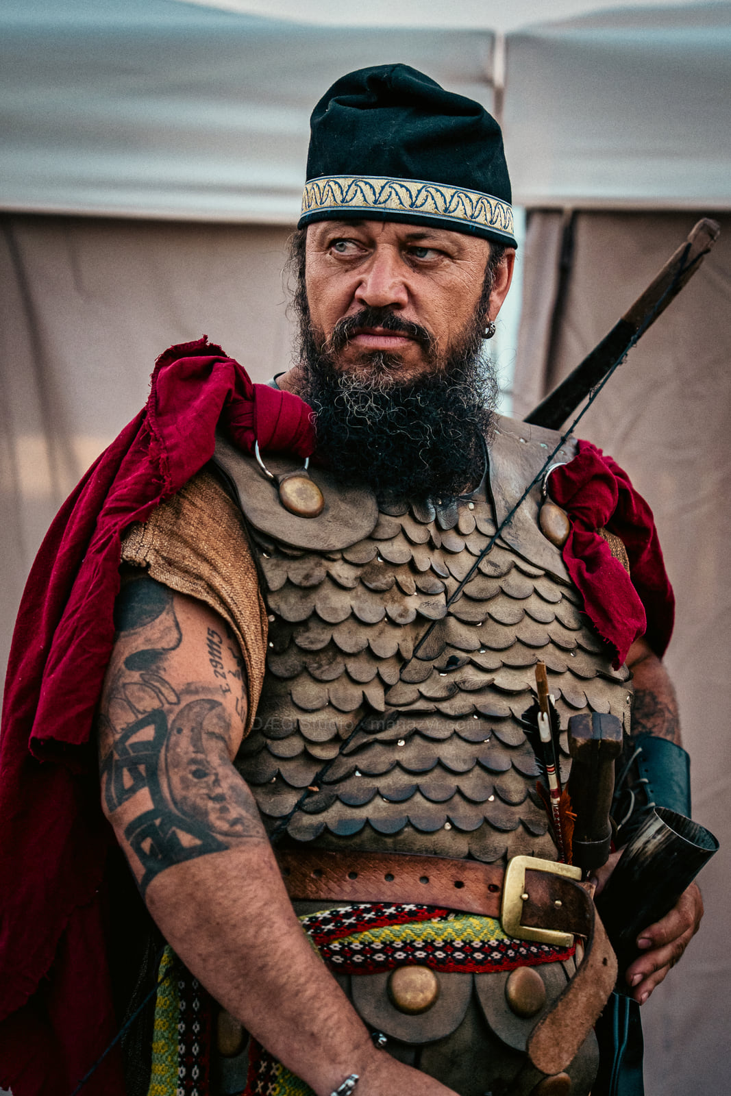 Tattooed reenactor in scale armor and a red cape stands ready outdoors.