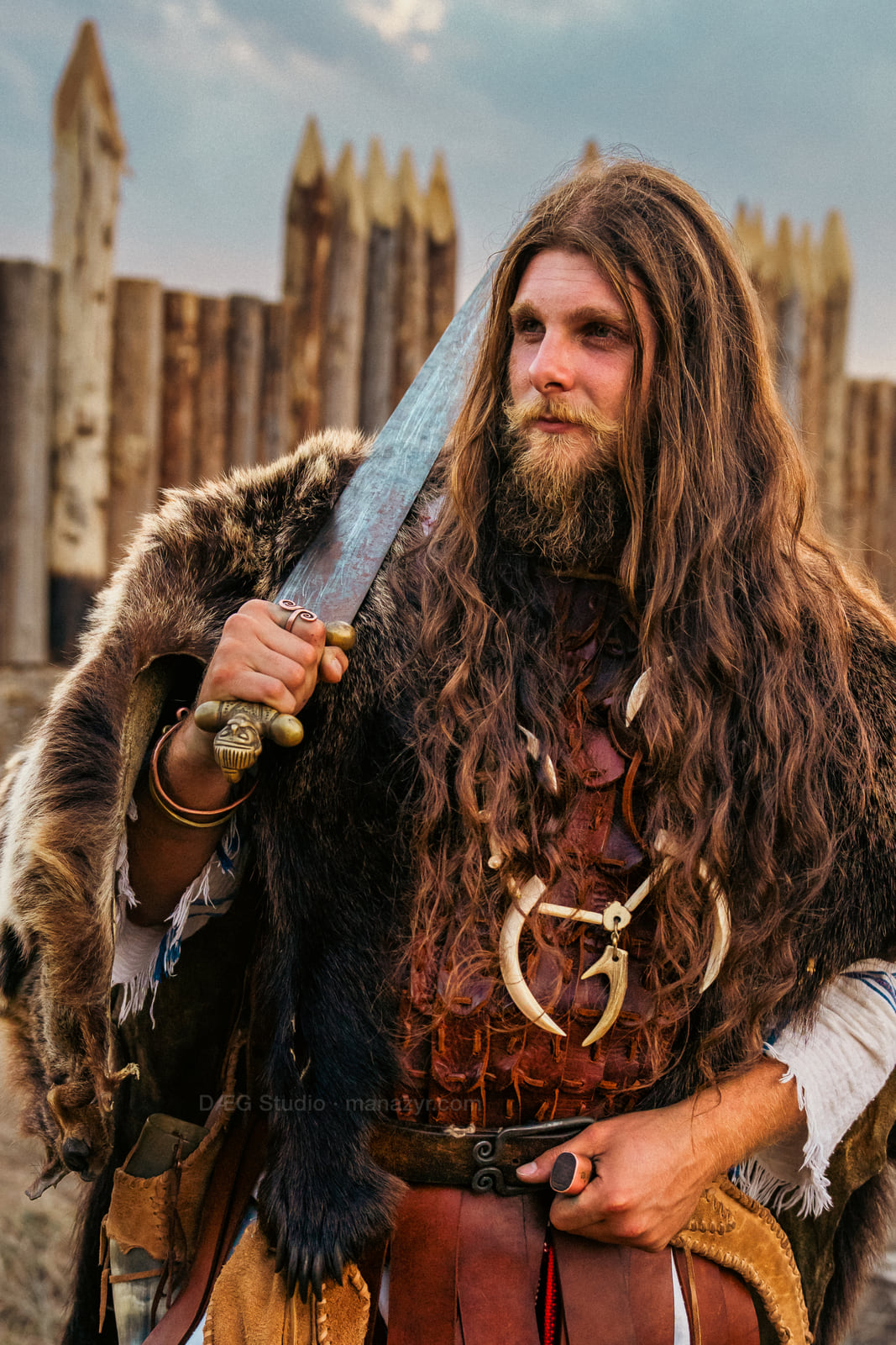 Long-haired reenactor in layered fur clothing holds a sword before a wooden palisade.