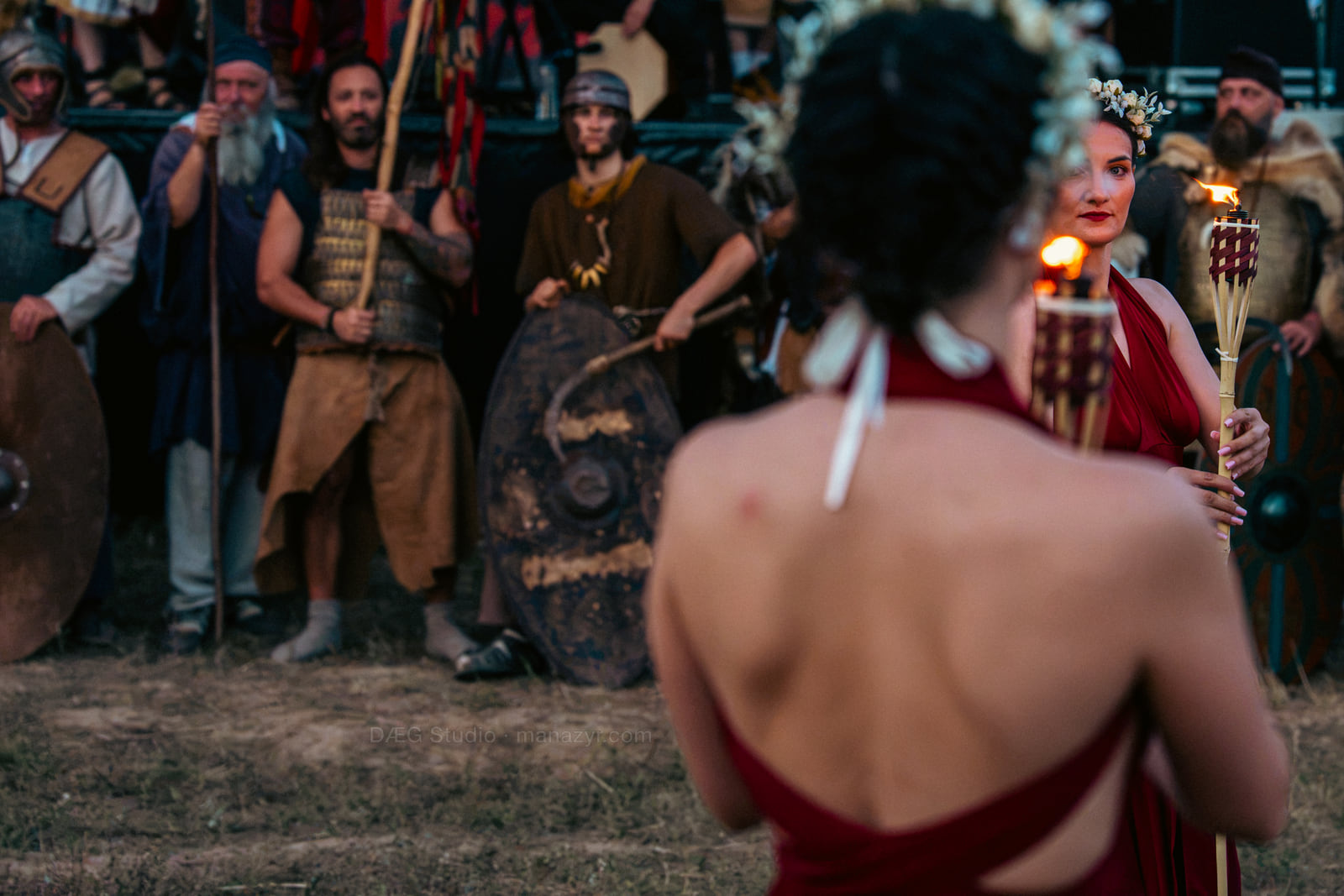 Two red-dressed performers carry flaming torches before a line of historical reenactors.