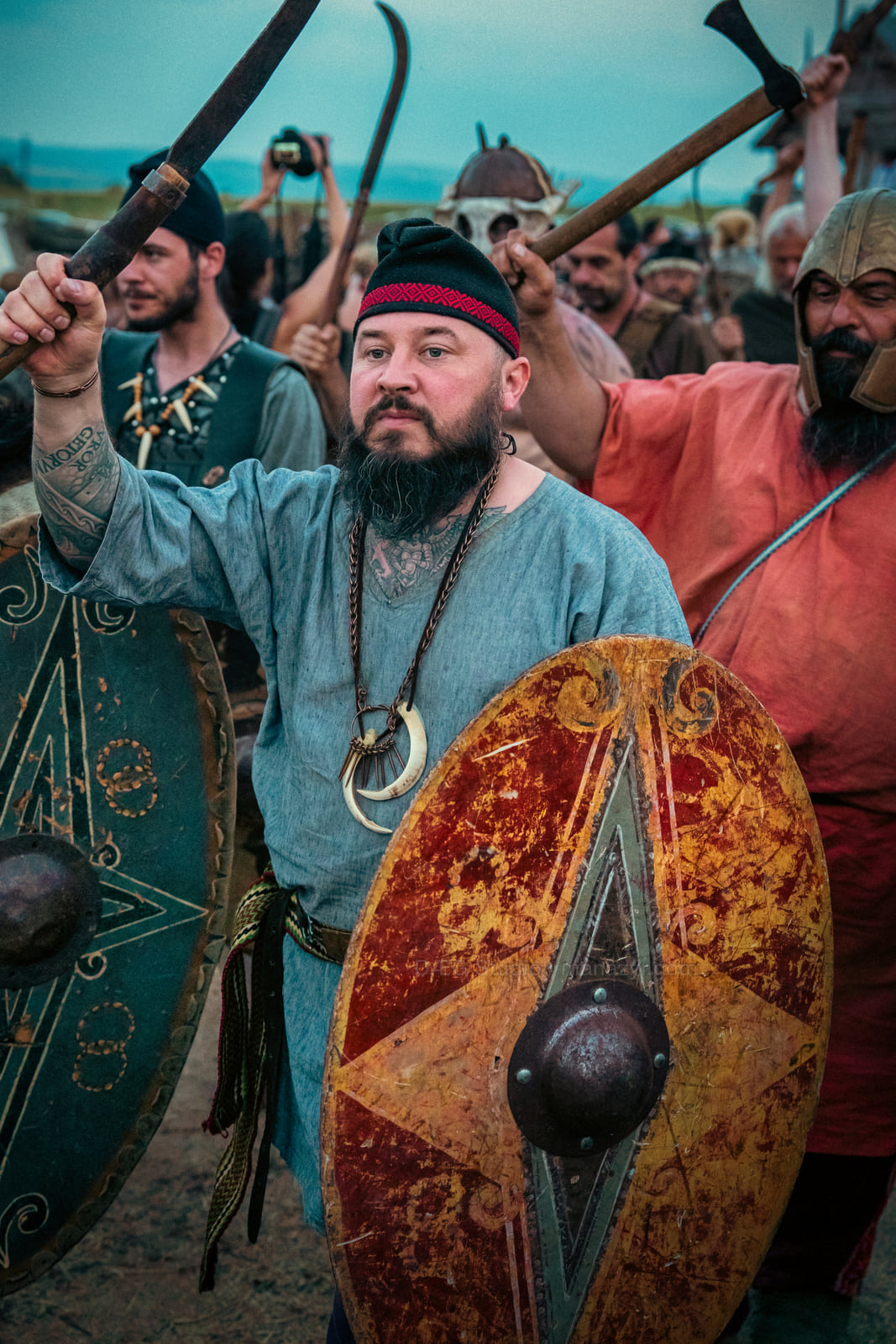 Bearded reenactor raises a weapon above a painted shield among an energized crowd.