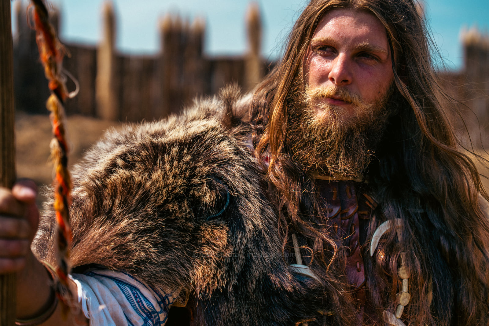 Long-haired reenactor in fur clothing holds a sword during an outdoor historical performance.