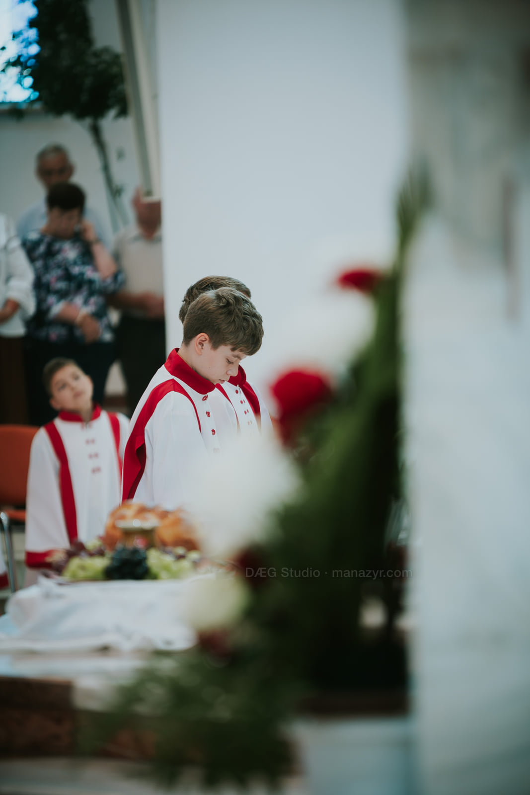 Un tânăr ministrant privește în jos lângă ofrande ceremoniale și flori estompate.