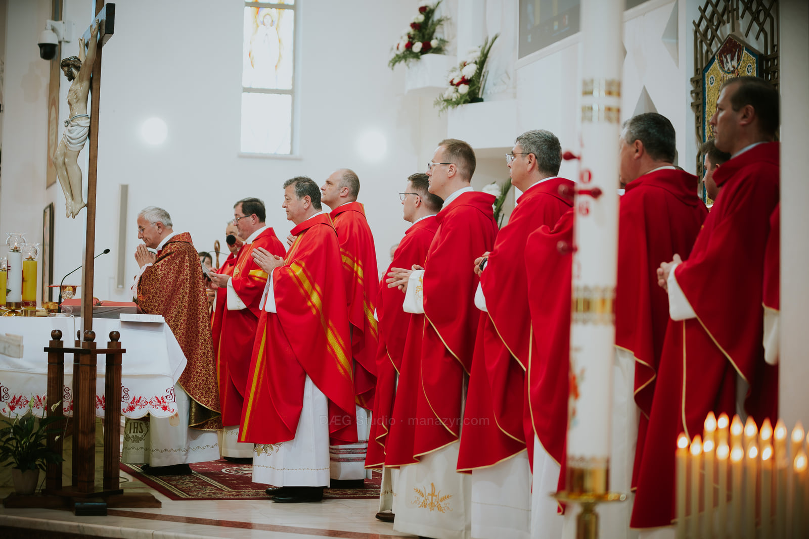 Un șir de clerici în veșminte roșii stă lângă altar și lumânările aprinse.