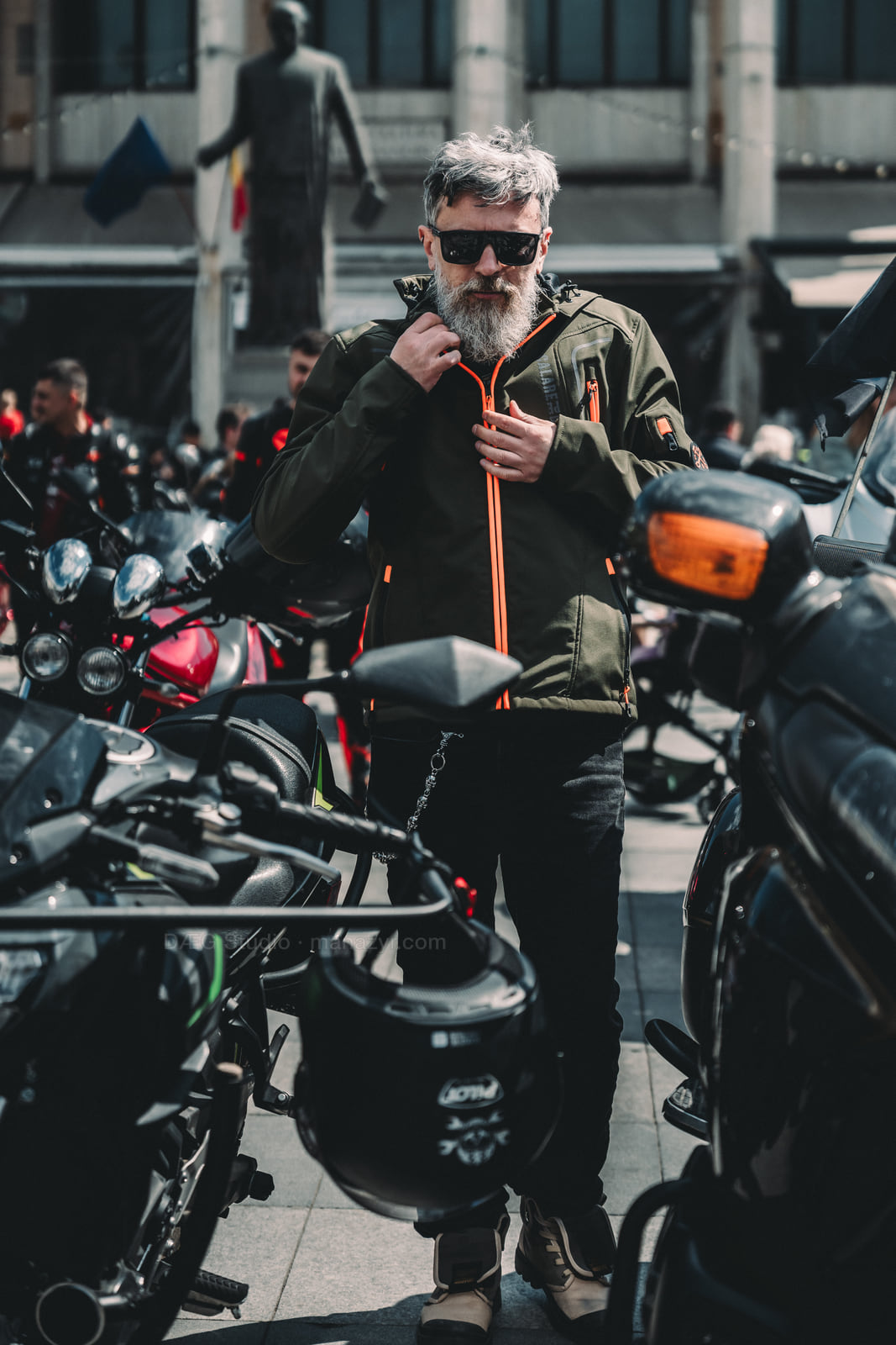 Bearded man adjusts his jacket while standing between parked motorcycles in sunlight.