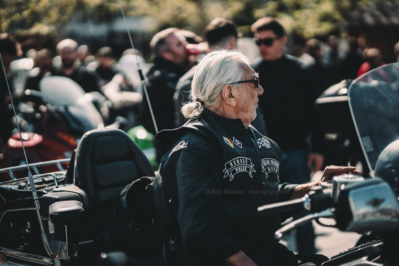 Older biker with tied-back white hair stands beside a touring motorcycle in the crowd.