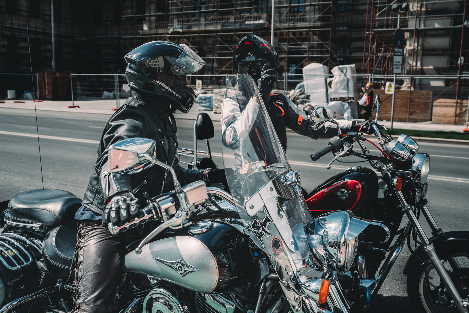 Two helmeted riders pause side by side among chrome motorcycles on a city street.