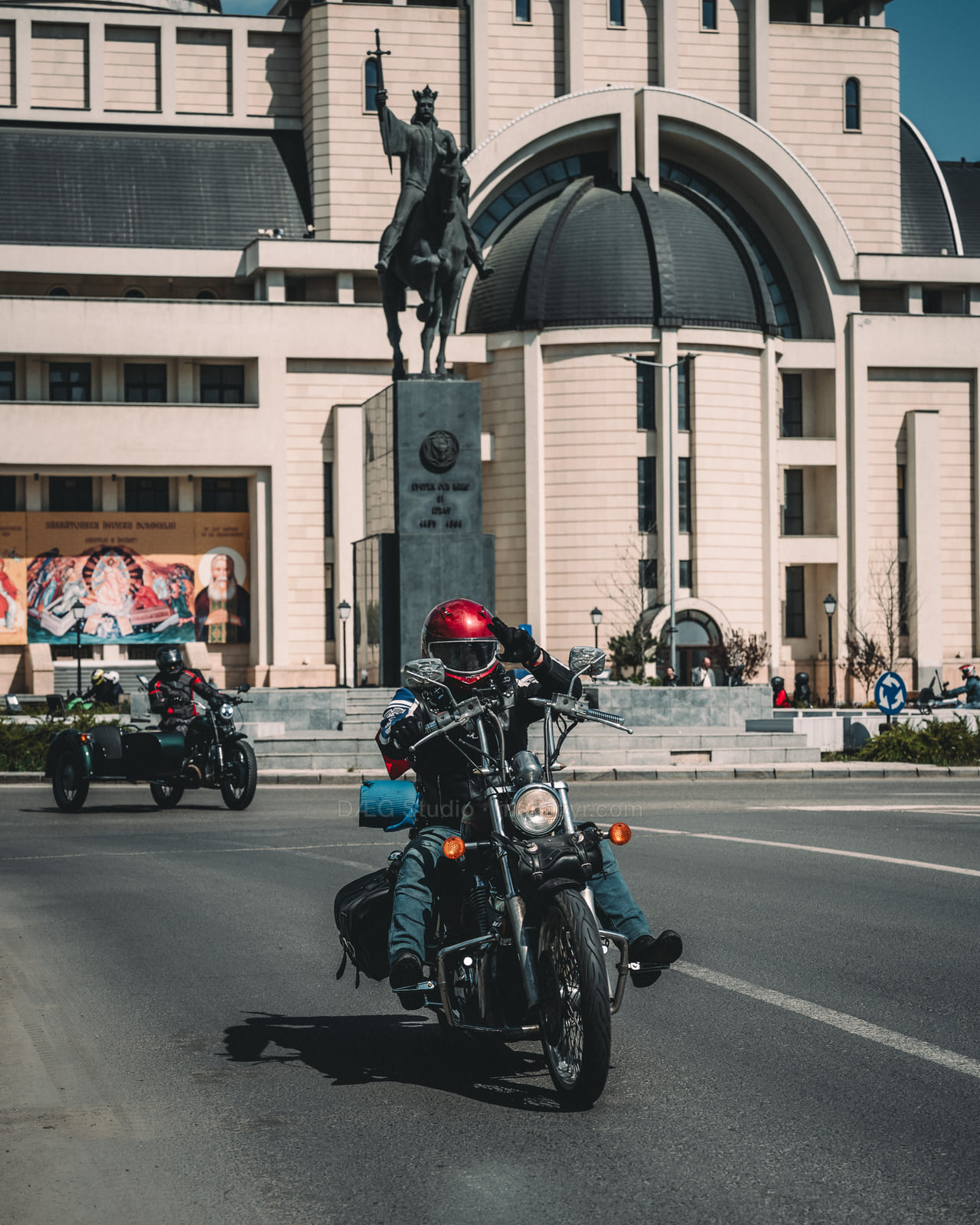 Helmeted rider salutes from a motorcycle before a large civic building and statue.