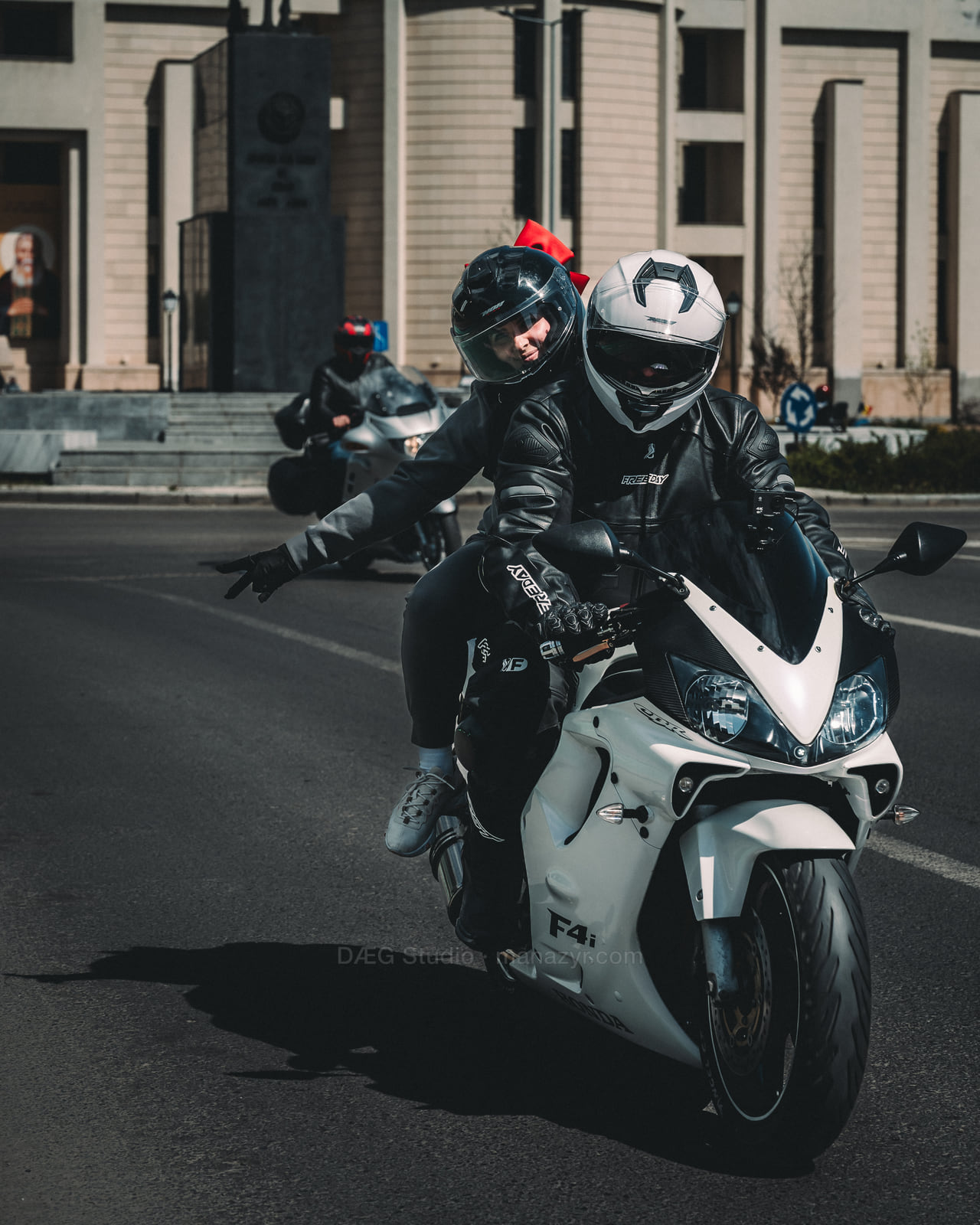 A passenger flashes a peace sign from behind the rider on a white sport motorcycle.